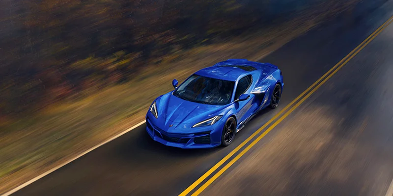 A top-down photo of a blue 2025 Chevy Corvette E-Ray driving down a wooded single-lane road near Clinton Township